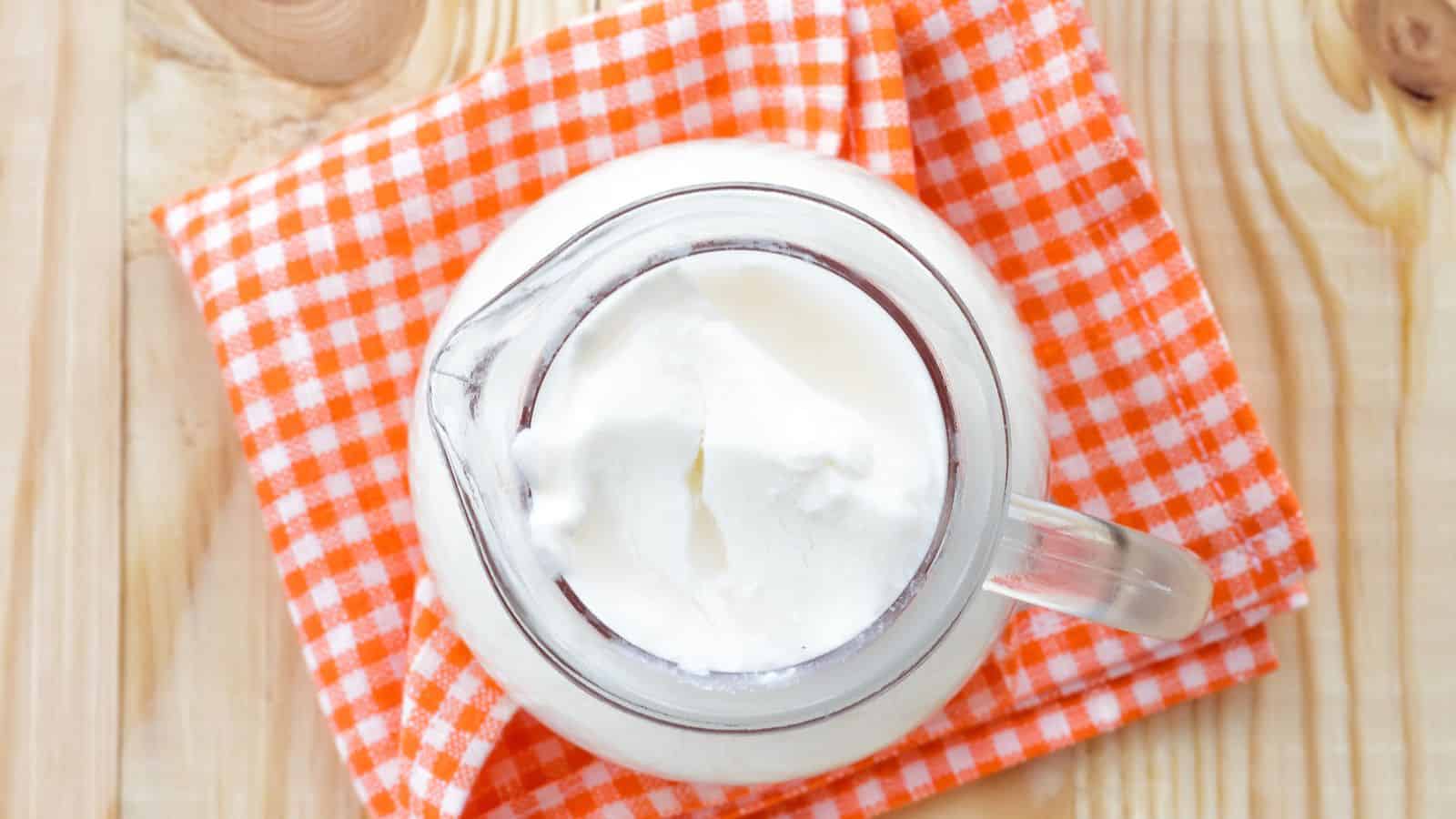 A clear glass pitcher filled with a thick white liquid, likely yogurt, is placed on an orange and white checkered cloth on a wooden surface. The photo is taken from above.