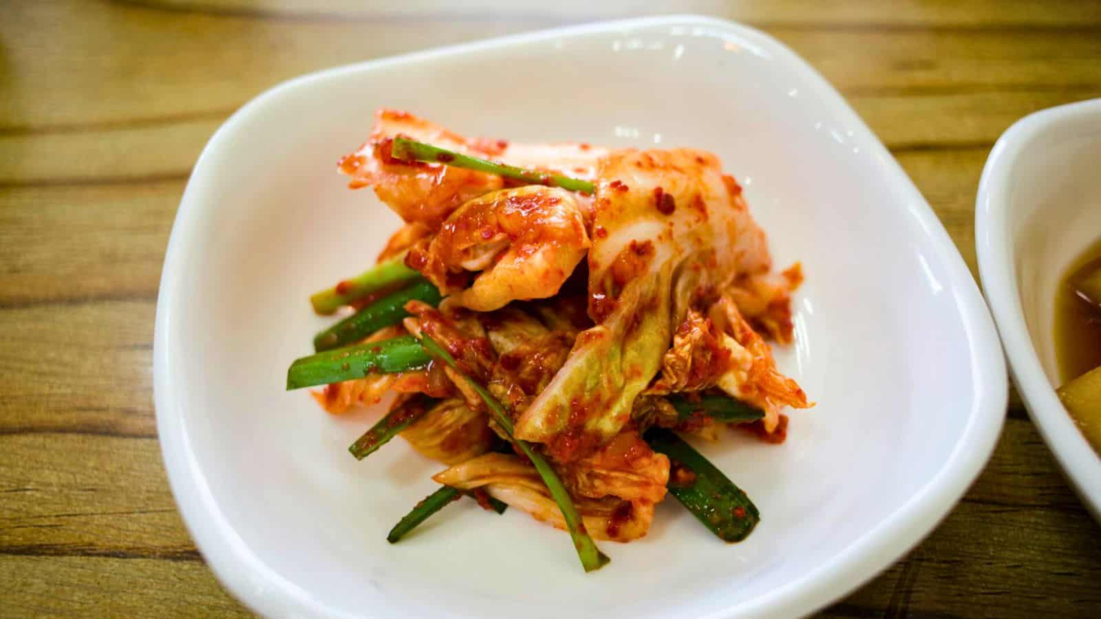 A plate of kimchi, a traditional Korean side dish made of fermented vegetables, primarily napa cabbage and radish, seasoned with chili pepper, garlic, ginger, and other spices, served in a white dish on a wooden table.