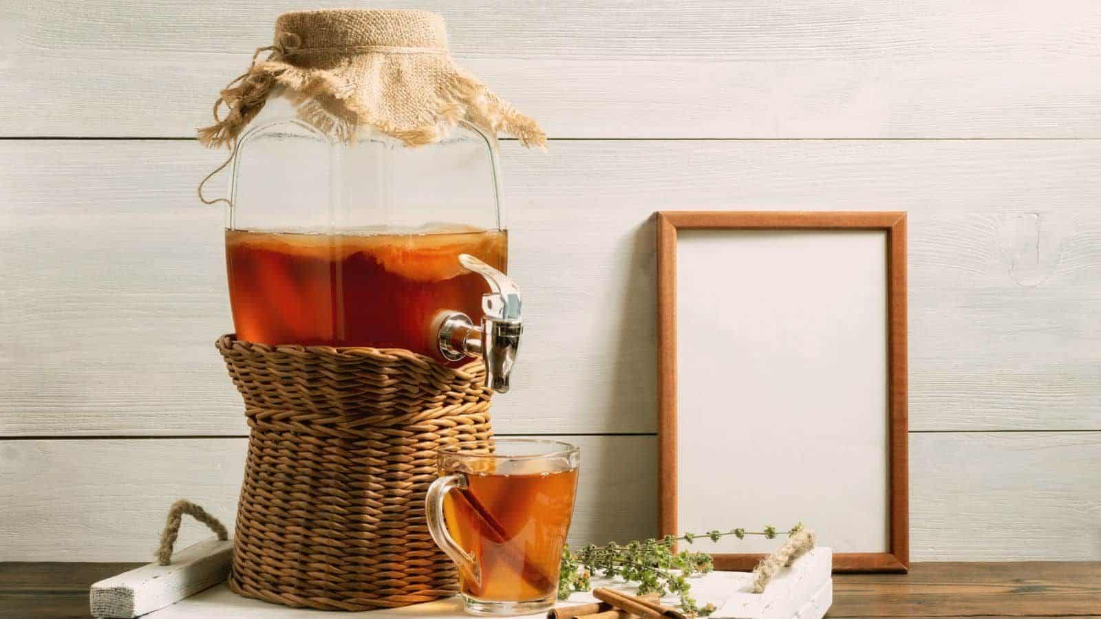 A glass beverage dispenser containing iced tea sits on a wicker stand. To the right, there is a blank wooden picture frame, and in front, there is a glass cup filled with iced tea. Green herbs and a cinnamon stick are placed beside the glass cup.