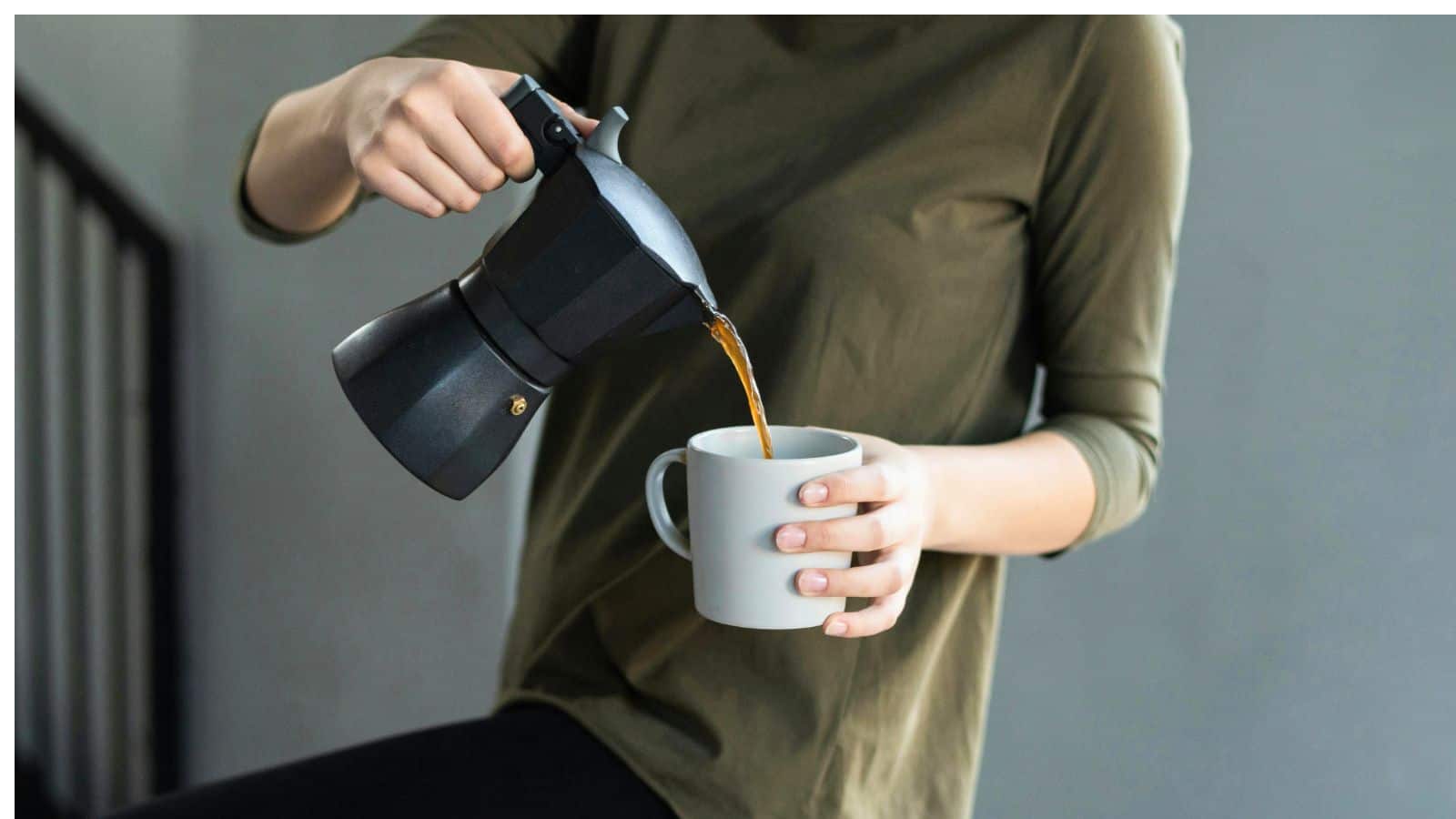 A person in a green shirt is pouring coffee from a black stovetop espresso maker into a white mug. The person is indoors, and part of a staircase is visible in the background.