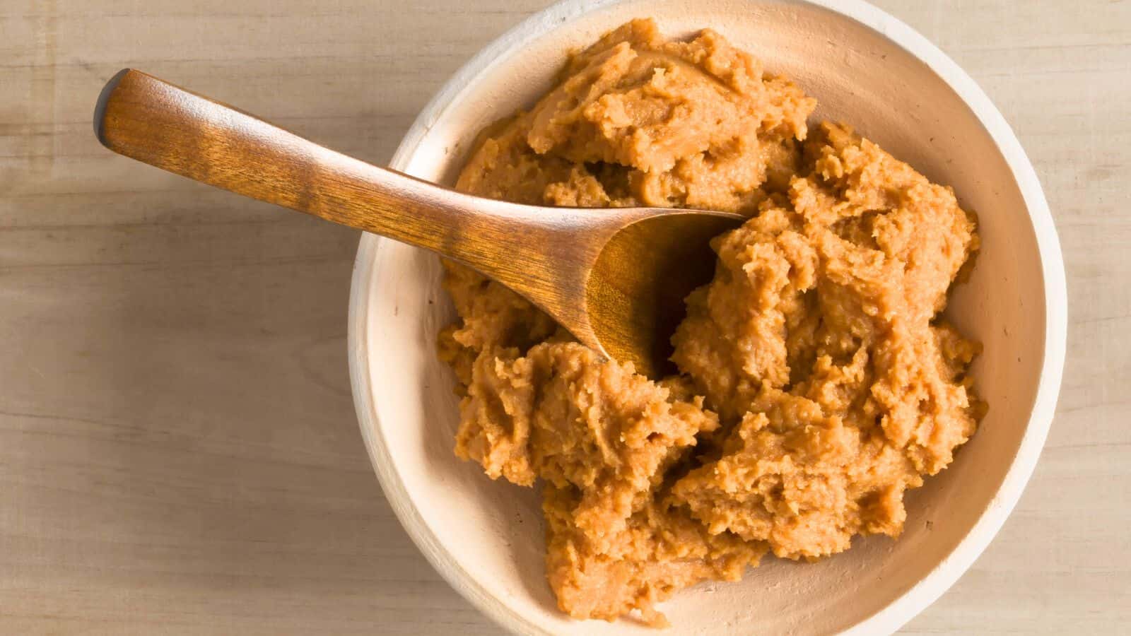 A wooden bowl filled with light brown miso paste. A wooden spoon is resting inside the bowl, partially submerged in the miso. The bowl is placed on a light-colored wooden surface.