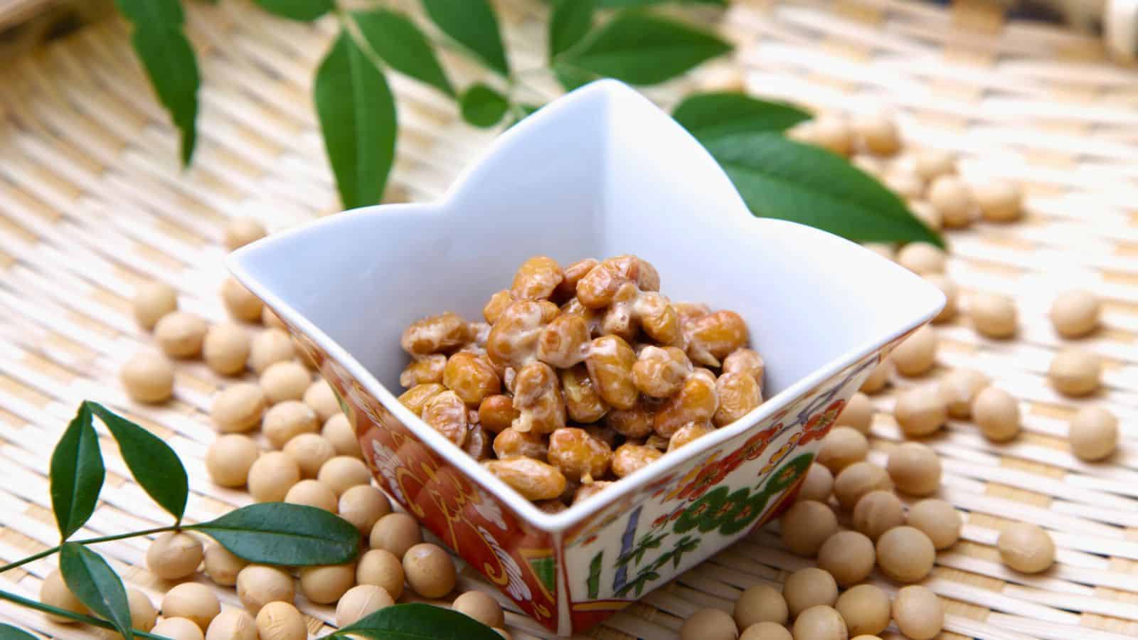 A square, ceramic bowl with intricate designs holds natto (fermented soybeans) on a woven mat. Soybeans and green leaves are scattered around the bowl.