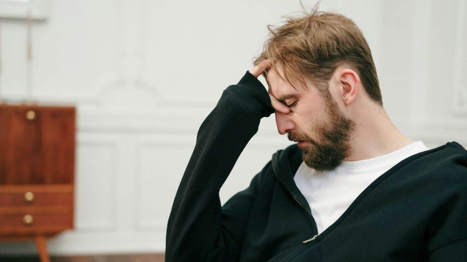 A man with a beard, wearing a white shirt and a black jacket, sits with his head resting on his hand. His eyes are closed and he appears to be in deep thought or tired. The background shows a simple interior with white paneled walls and a wooden piece of furniture.