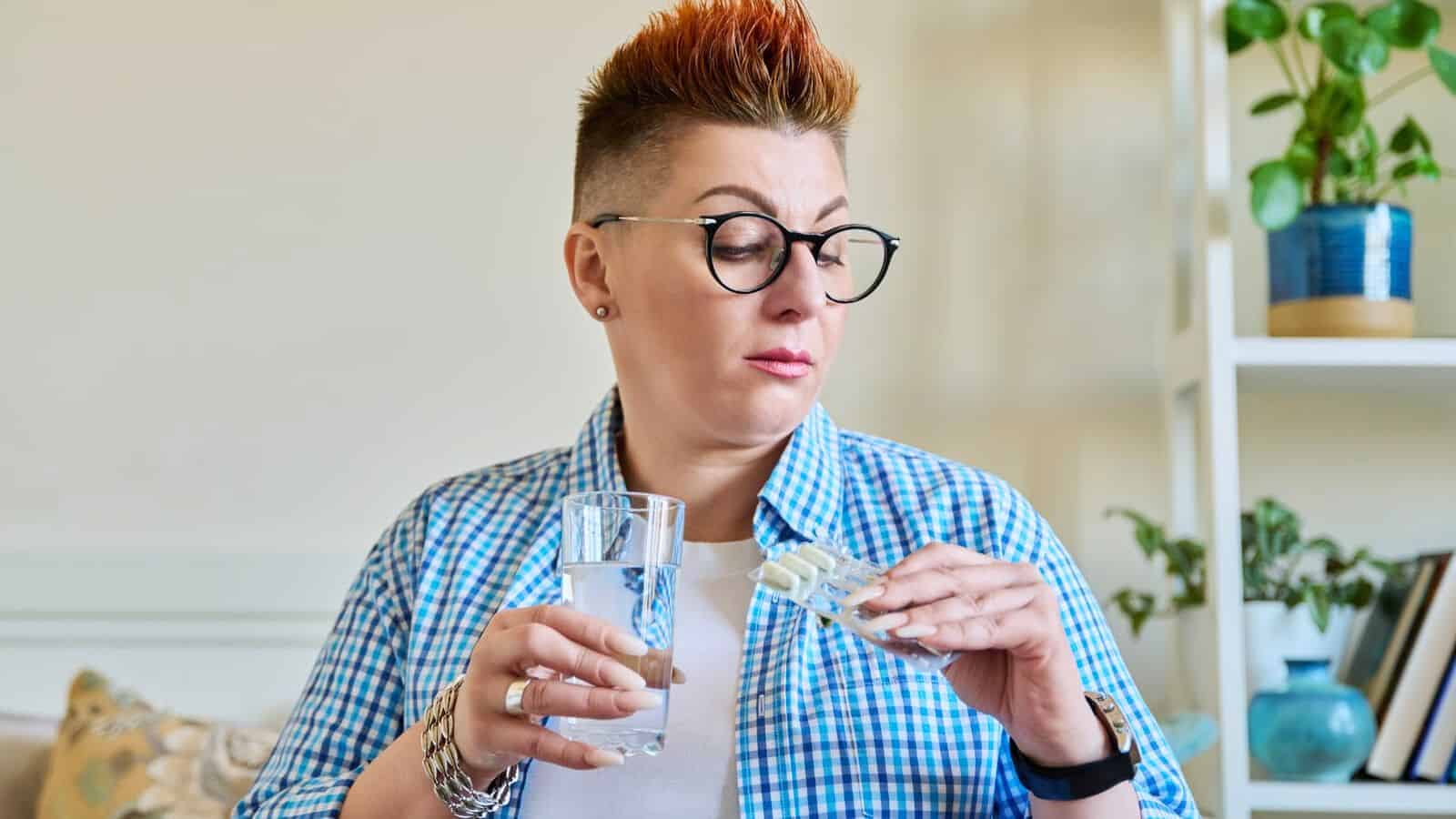 A person with short, styled hair and glasses is sitting in a room. They are holding a glass of water in one hand and a package of tablets in the other, appearing to read the label. They are dressed in a checkered shirt. A bookshelf and plant are visible in the background.