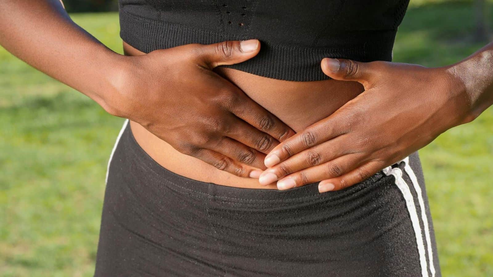 A person wearing a black crop top and black pants with white stripes places their hands on their stomach. The person is standing outside, and green grass can be seen in the background.