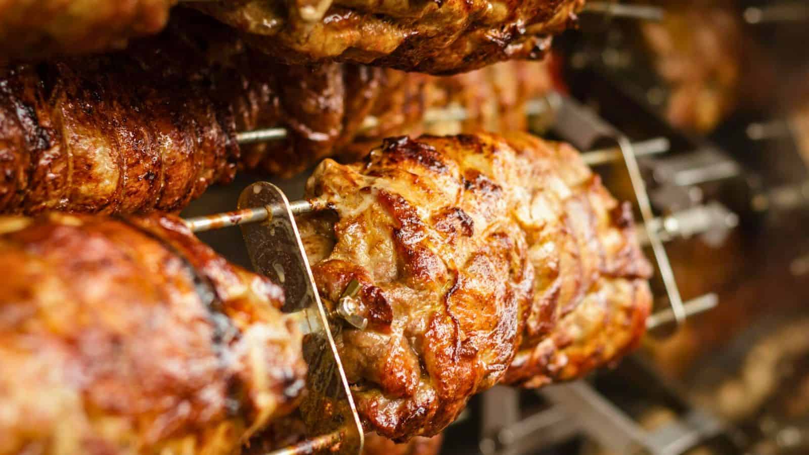 Close-up image of seasoned meats roasting on rotating skewers in a commercial kitchen. The golden-brown, crispy exterior of the meat indicates it is well-cooked. The setup suggests it is a rotisserie-style preparation.