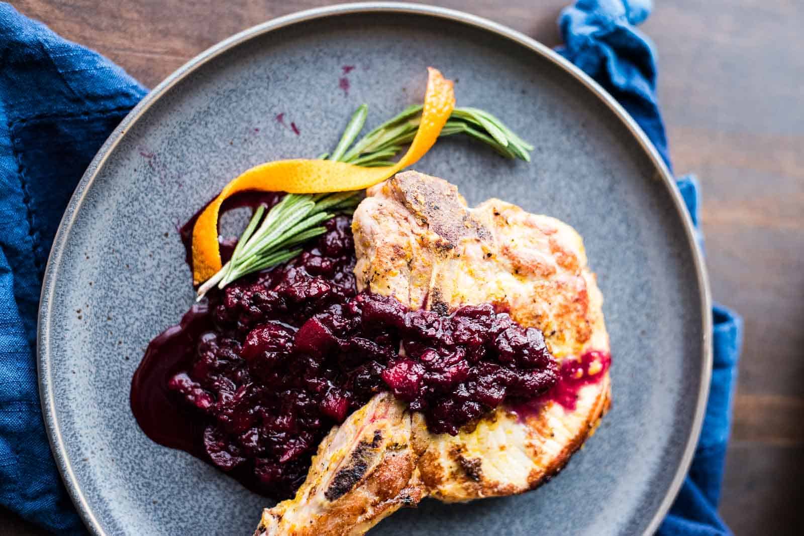 A grilled piece of meat, garnished with a rosemary sprig and a strip of orange peel, is served on a gray plate with a dark red sauce.