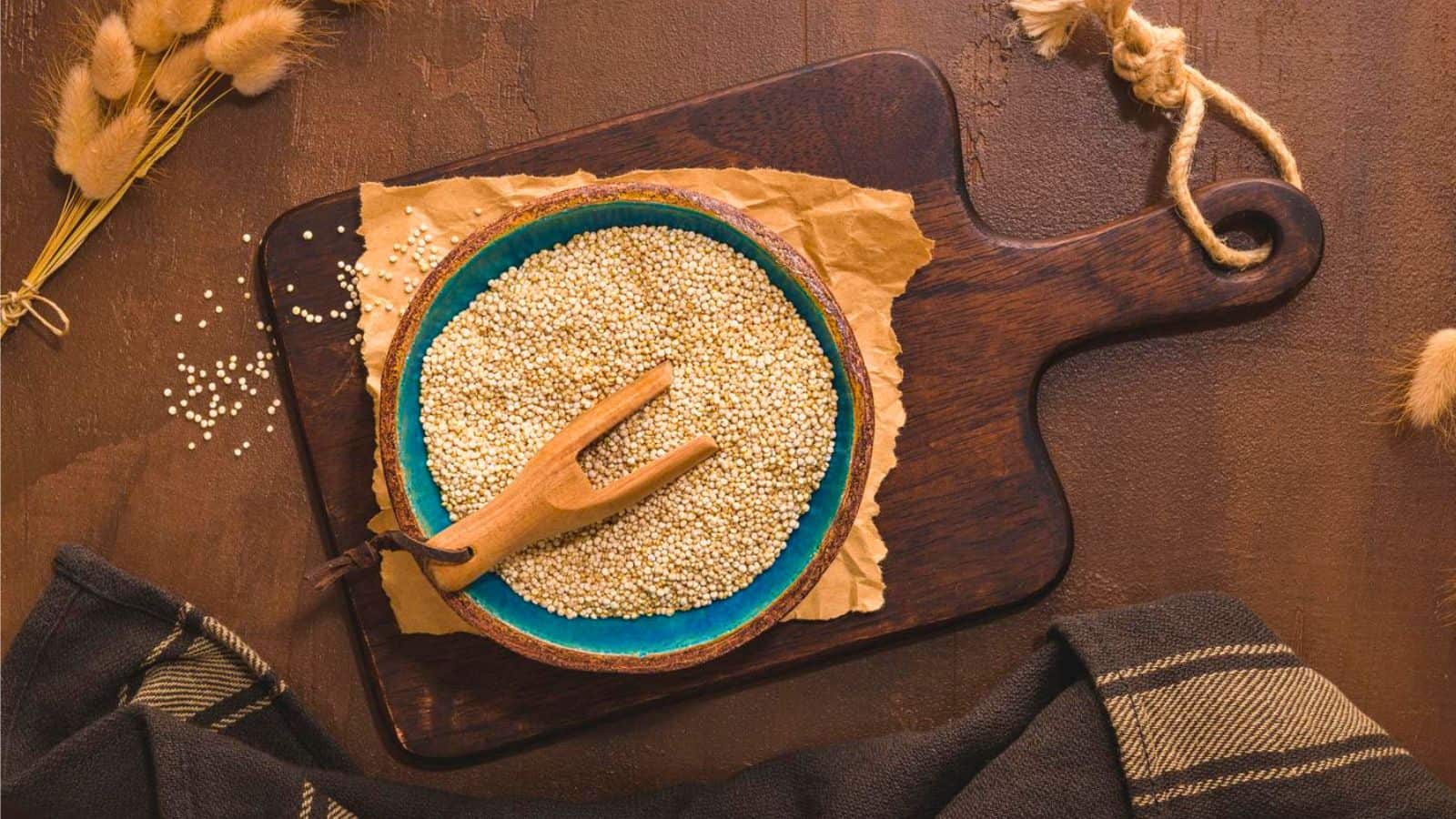 A bowl filled with quinoa sits on a piece of parchment paper on a dark wooden cutting board. A small wooden fork is in the bowl, and decorative wheat stalks and a dark cloth with stripes are placed around the board.