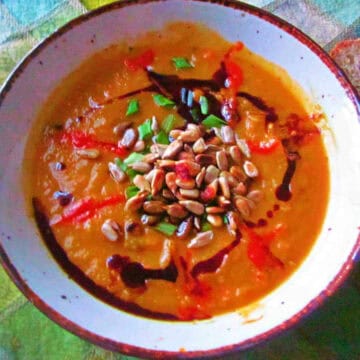 A bowl of soup garnished with seeds, chopped herbs, and drizzles of sauce. The bowl is placed on a checkered green cloth beside a spoon and a slice of bread, reminiscent of the famous dishes from the 60s.