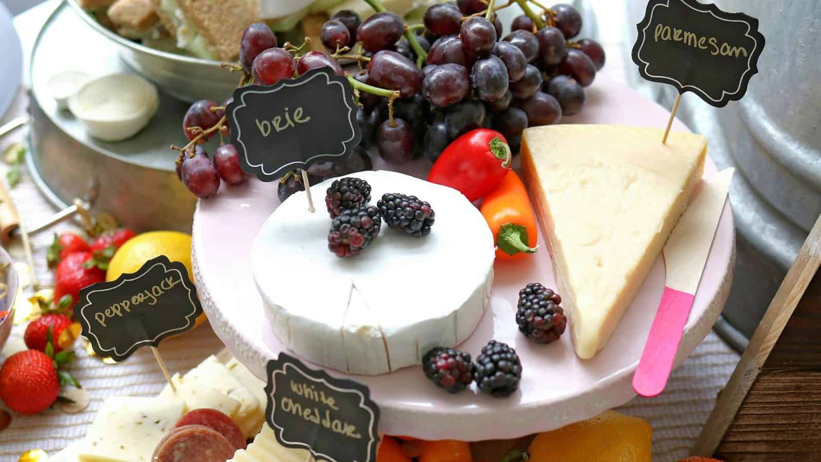 A platter featuring various cheeses, including a wedge of Parmesan, a round of brie topped with blackberries, and white cheddar. The platter is garnished with grapes, strawberries, bell peppers, and lemon slices. Small chalkboard signs label the items.