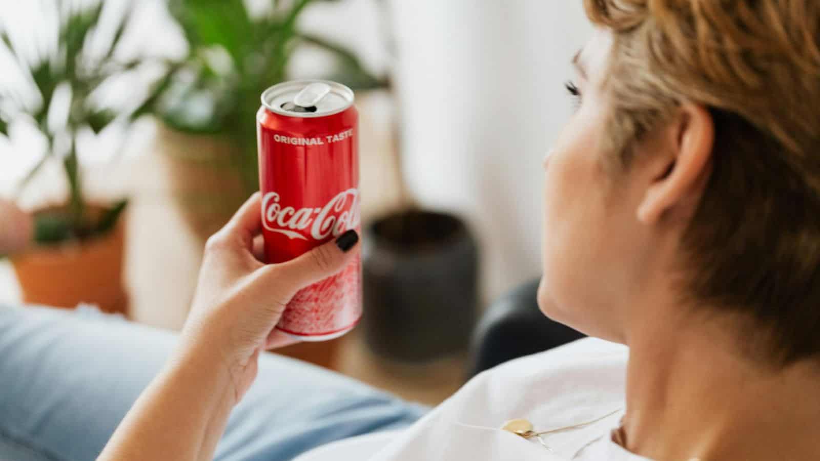 A person holding a can of Coca-Cola with the label "Original Taste." The individual is shown from behind and slightly to the side, with a focus on the drink. There are potted plants in the blurred background.