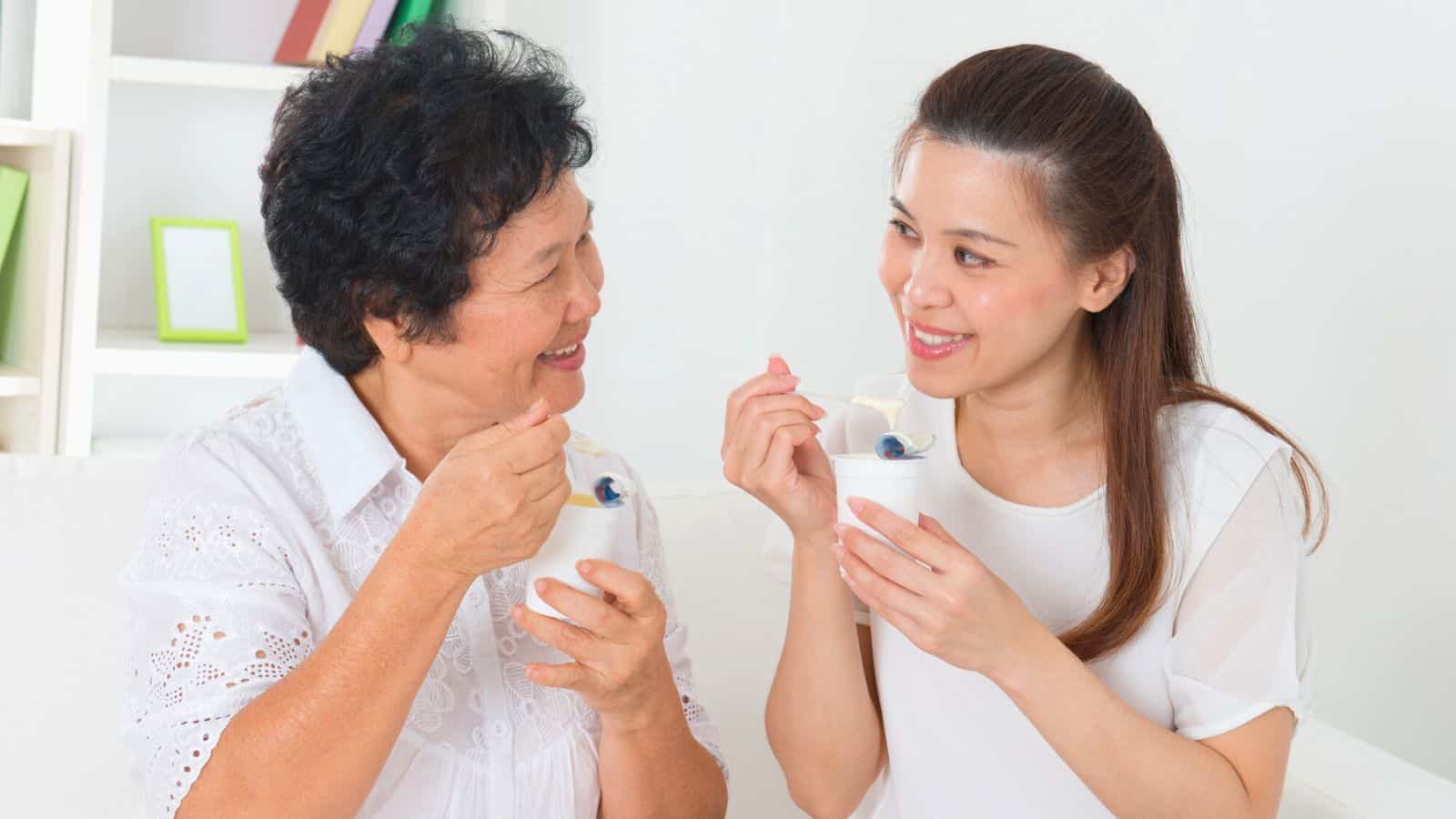 Two women are sitting together, smiling, and eating yogurt. The older woman on the left has short black hair and is wearing a white blouse, while the younger woman on the right has long brown hair tied back and is also wearing a white blouse.