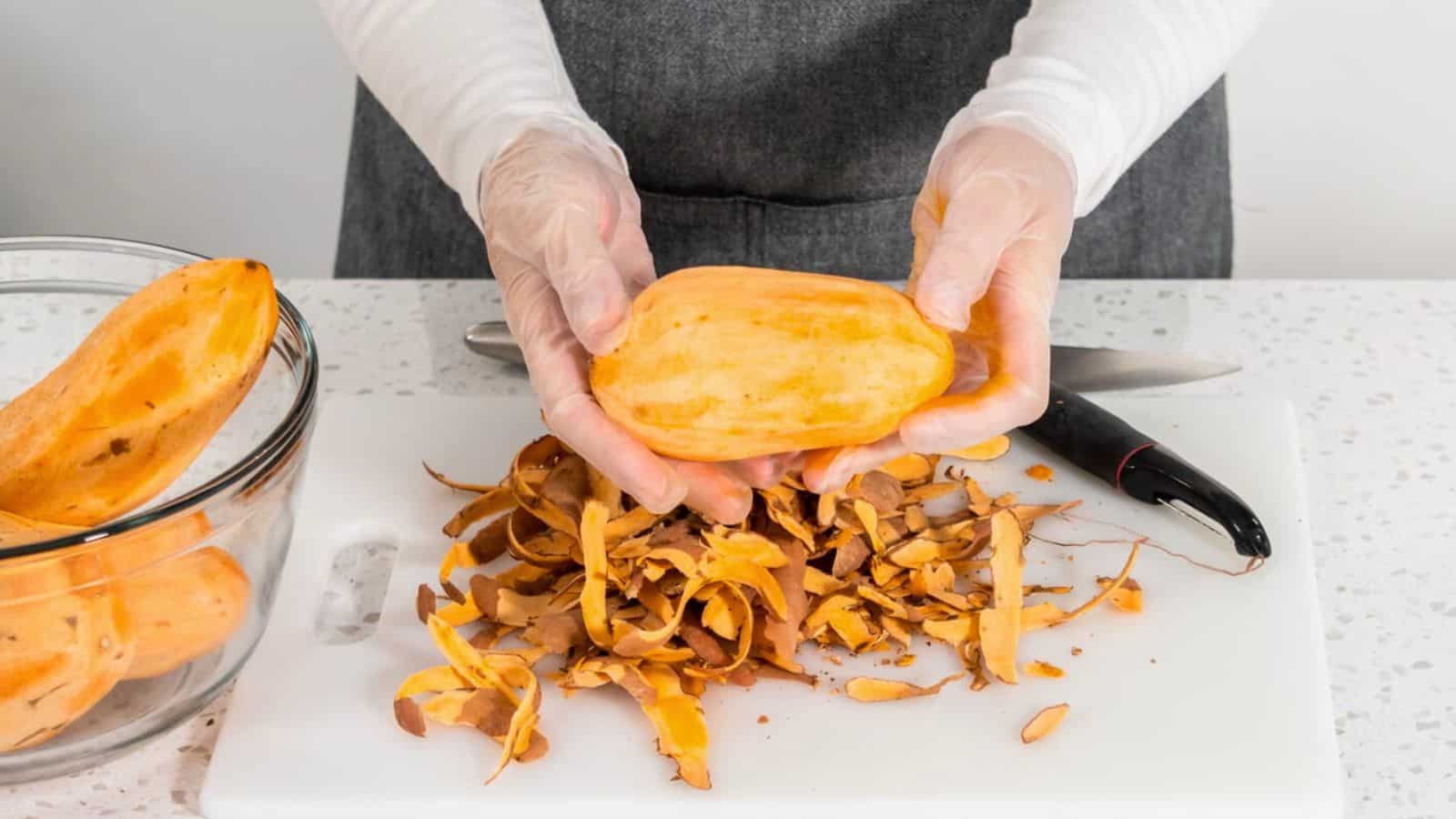 A person wearing gloves is peeling a sweet potato over a white cutting board. There are peeled sweet potato skins and a black-handled knife on the board. A bowl containing other sweet potatoes is on the left side of the cutting board.
