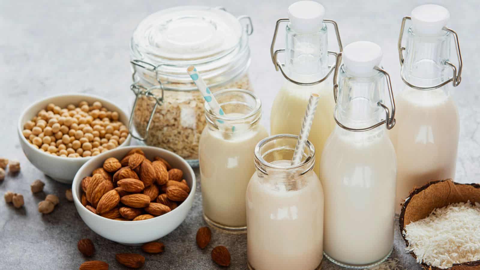 Various plant-based milk alternatives and their ingredients are displayed on a table. There are glass bottles and jars filled with almond milk, soy milk, and oat milk. Bowls containing soybeans, almonds, oats, and coconut pieces are also shown.