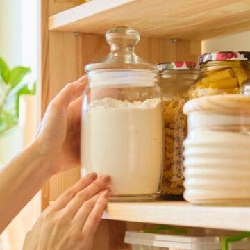 A person is placing a large glass jar filled with white powder, likely flour, on a wooden kitchen shelf. The shelf also holds various other jars with different dry goods. There are green plants on the windowsill in the background.