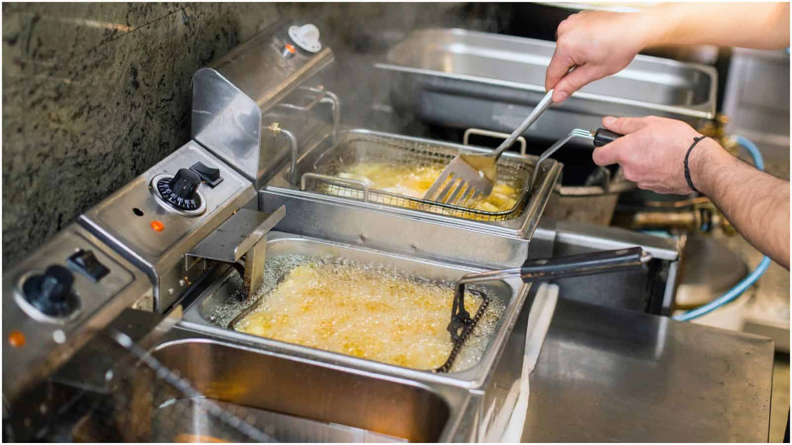 A person is using metal tongs to fry food in a deep fryer. The fryer contains hot oil, visibly bubbling. The person is cooking in a commercial kitchen setting. Multiple fryers and stainless steel counters are shown in the background.