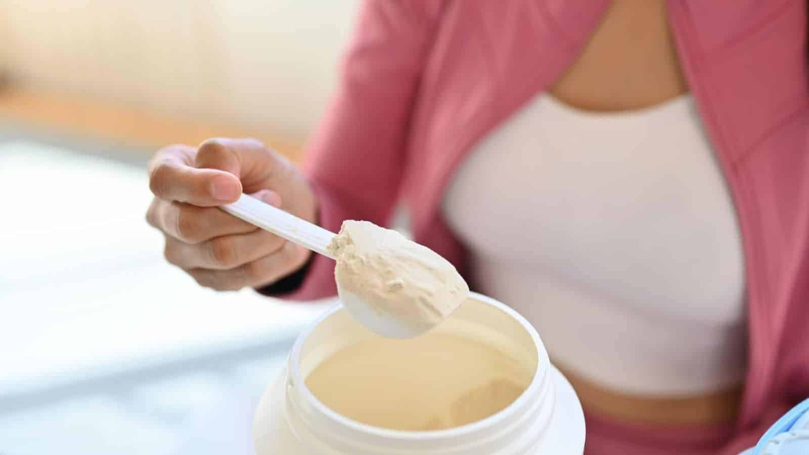 A person in a pink jacket and white top is scooping powder from a large white container using a plastic scoop. The container appears to be filled with a light, beige-colored powder.