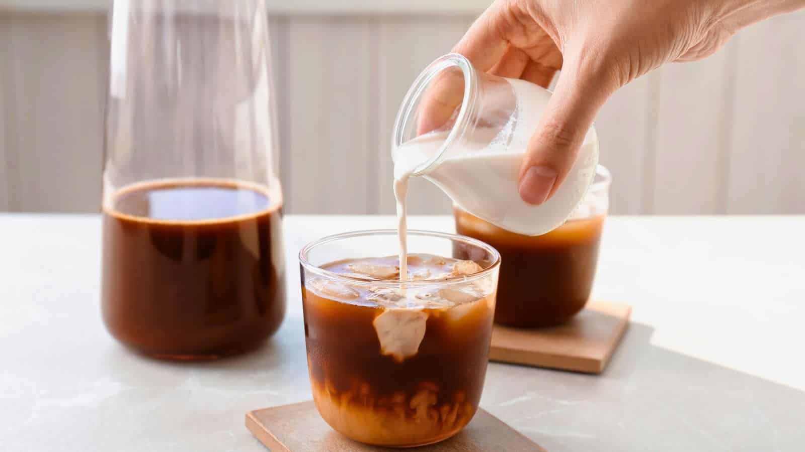 A hand is pouring milk from a small glass jar into a glass of iced coffee. Another glass of iced coffee and a larger glass container filled with black coffee are placed on wooden coasters nearby.