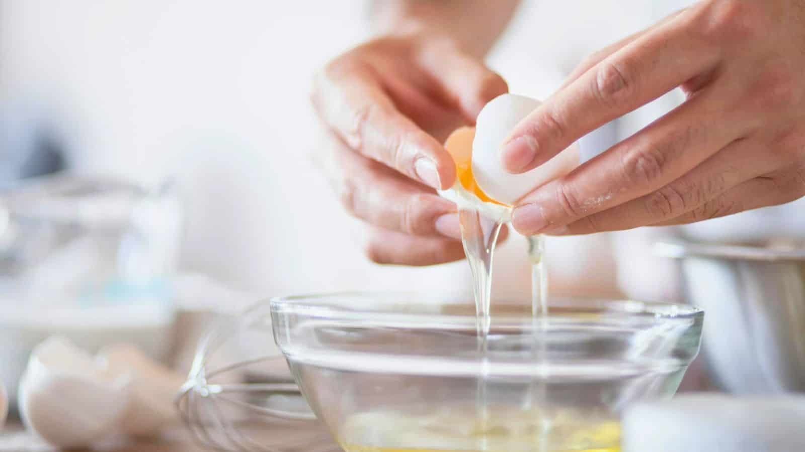 Close-up of hands cracking an egg into a glass bowl. The egg yolk and white are falling into the bowl, while another cracked egg is visible in the background along with blurry kitchen utensils.