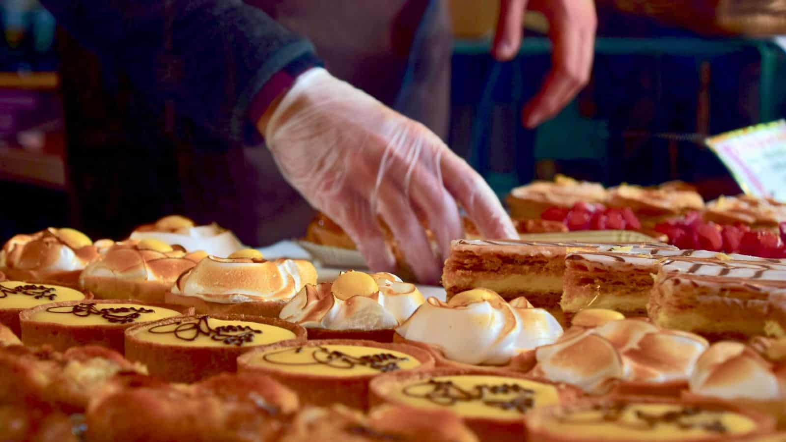 A person wearing a glove reaches out to select a dessert from a variety of pastries. The assortment includes lemon meringue tarts, rectangular layered cakes with icing, and other decorated sweets displayed on a table.