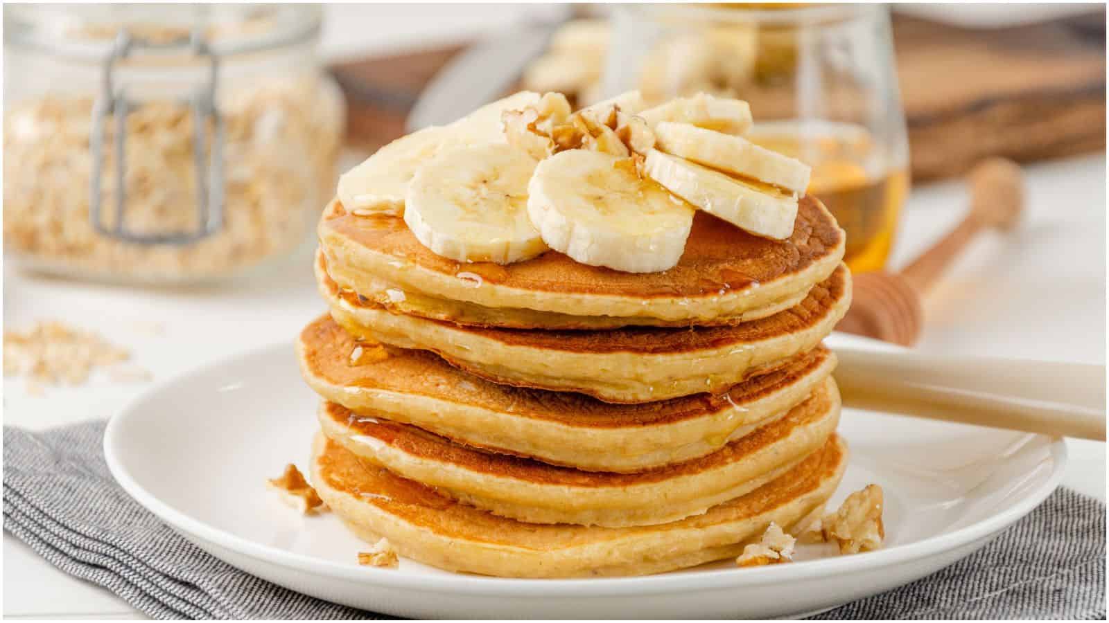 A stack of pancakes topped with banana slices, chopped nuts, and a drizzle of honey sits on a white plate. The plate is placed on a gray napkin, and there are oats and a jar of honey in the background.