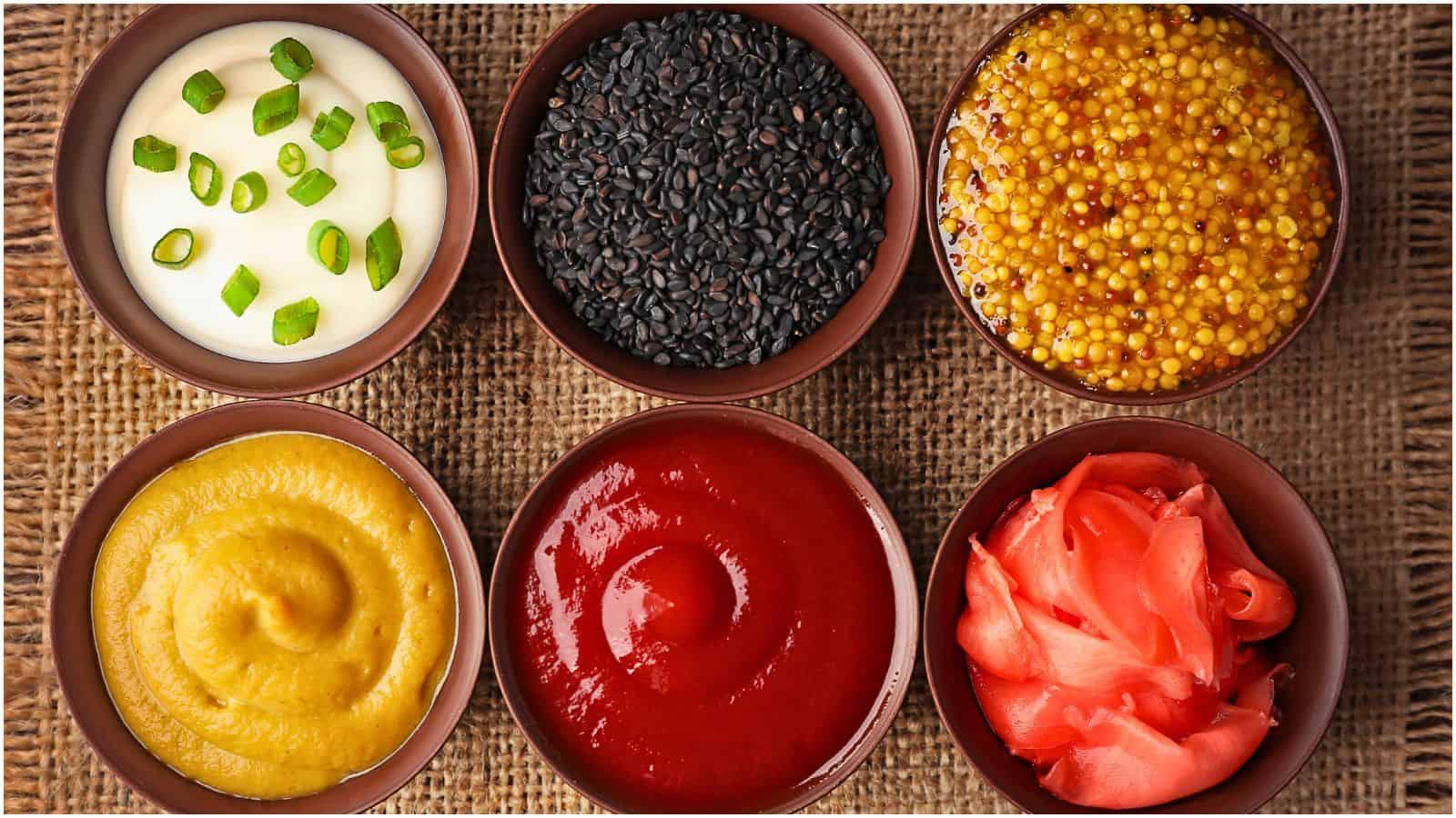 Six bowls of food arranged in two rows. The top row contains chopped green onions and white sauce, black sesame seeds, and whole grain mustard. The bottom row contains yellow mustard, ketchup, and pink pickled ginger. The bowls are on a burlap surface.