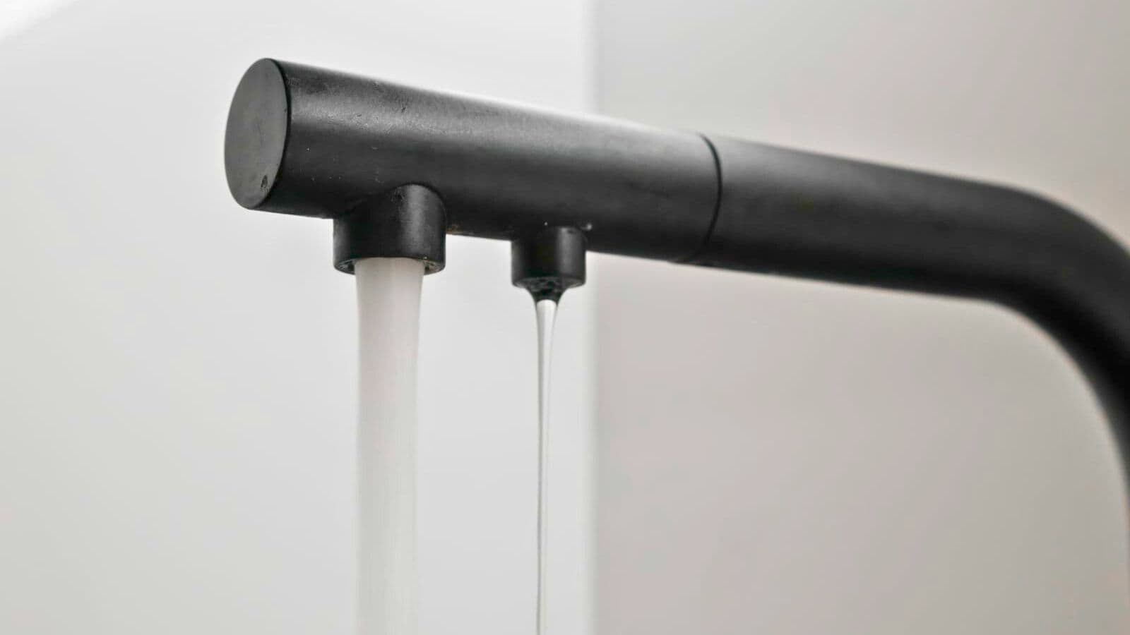A close-up image of a black faucet with two separate spouts. The left spout is dispensing water in a steady stream, while the right spout has a slow trickle of water. The background is out of focus and light-colored.