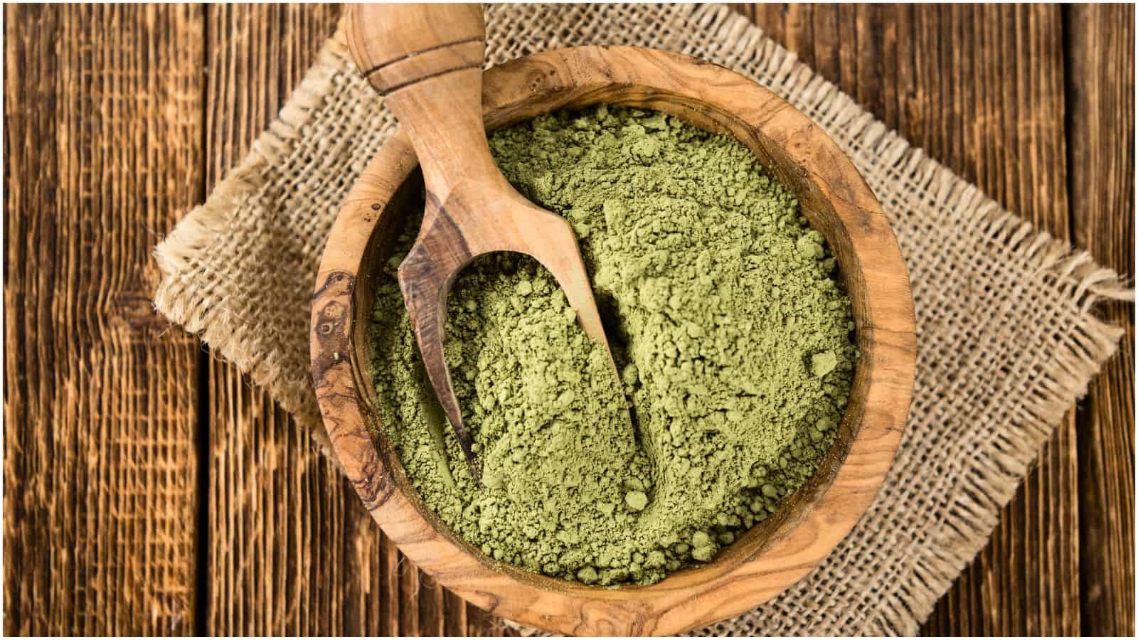 A wooden bowl filled with green powder sits on a piece of burlap fabric on a rustic wooden table. A wooden scoop is placed in the bowl, partially immersed in the powder. The setting has a natural, earthy feel with the materials and tones used.