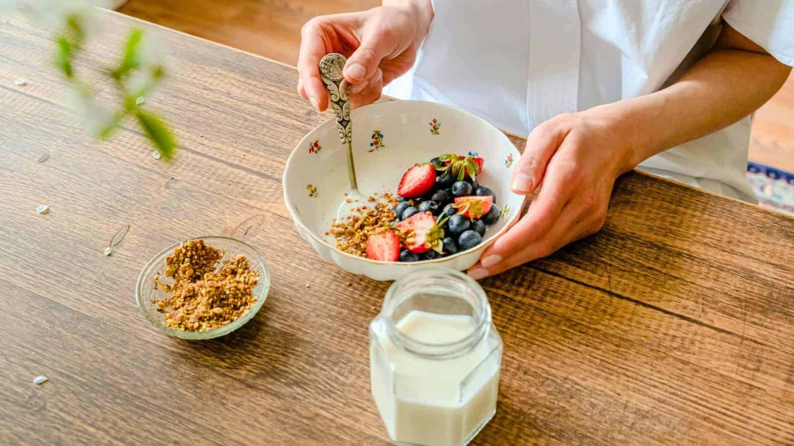 A person stirs a bowl of cereal topped with strawberries and blueberries using a spoon. A small bowl of seeds and a jar of milk are placed on the wooden table next to the bowl of cereal. The person is dressed in a white shirt.