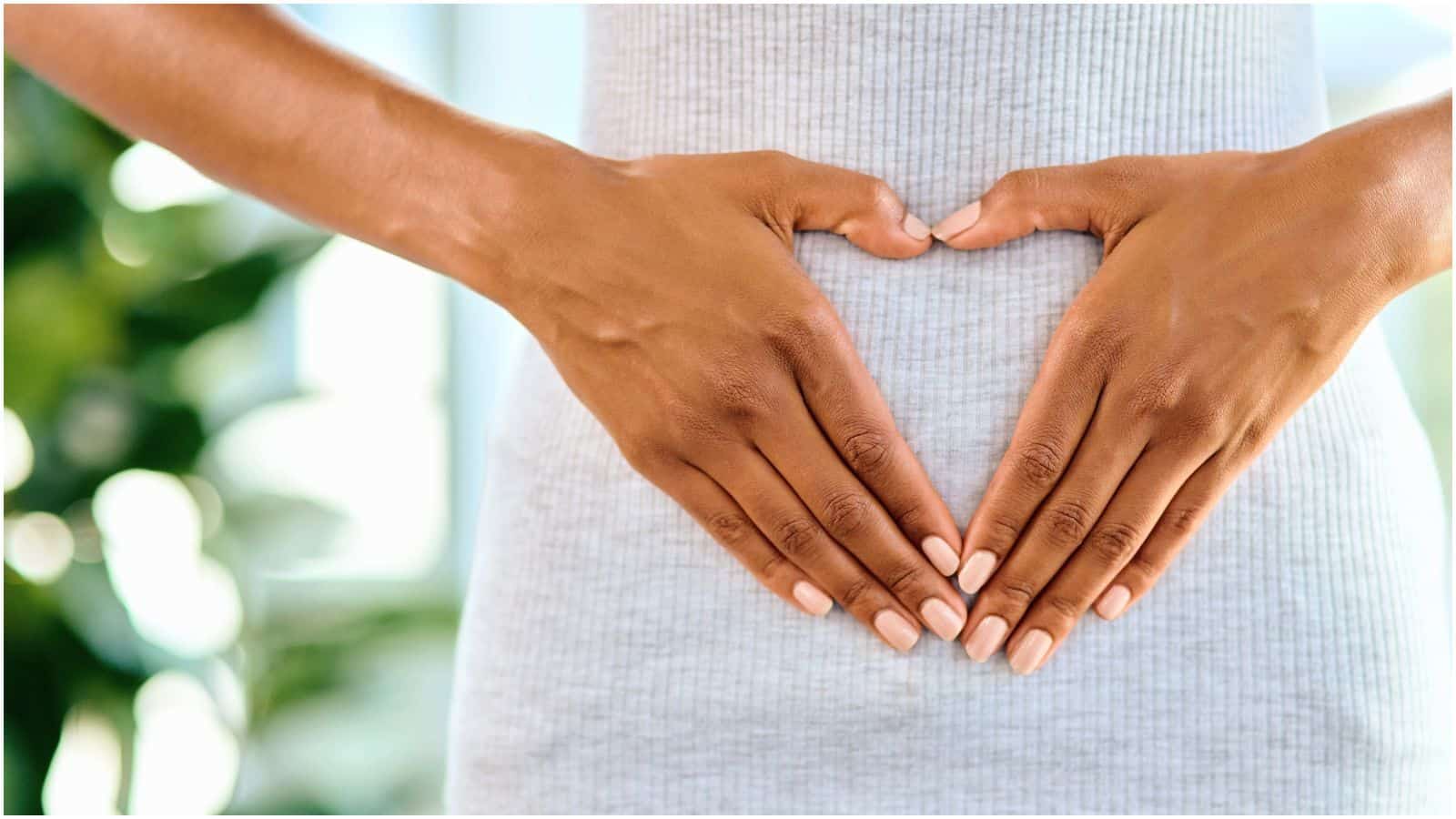 Close-up of a person forming a heart shape with their hands over their stomach. The person is wearing a light grey, ribbed top, and their nails are neatly manicured with a nude polish. The background is softly blurred with greenery.
