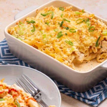 A baked pasta dish with golden cheese and green onion garnish sits in a rectangular casserole dish, reminiscent of Grandma's classic casseroles. A blue-patterned cloth lies beneath, evoking cozy Monday dinners. A serving rests on a gray plate next to a fork, with a small bowl of sliced green onions in the background.