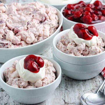 Bowls of creamy dessert topped with cherries and whipped cream are arranged on a wooden surface, perfect for potluck recipes. A red checkered cloth and three spoons are placed nearby, inviting you to indulge in these loveable recipes.