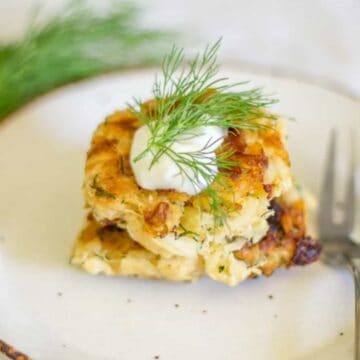 A small portion of a savory dish is served on a white plate. The dish appears to be a baked zucchini patty topped with a dollop of white sauce and garnished with a sprig of fresh dill, perfect for those exploring zucchini recipes. A fork is placed beside the food on the plate.