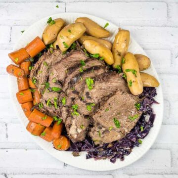 A white oval plate contains sliced pot roast, surrounded by roasted carrots, fingerling potatoes, and a bed of shredded red cabbage. The dish, perfect for slow cooker meals, is garnished with chopped parsley. The background is a light, textured surface.