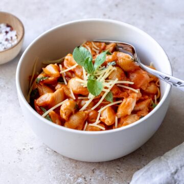 A white bowl filled with pasta in a red sauce, garnished with grated cheese and a sprig of basil. A spoon is placed inside the bowl. An Auto Draft small wooden bowl with a white seasoning is in the background on the left side. A light-colored napkin is on the lower right.