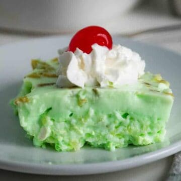 A slice of green gelatin dessert topped with a dollop of whipped cream and a red cherry is served on a white plate.