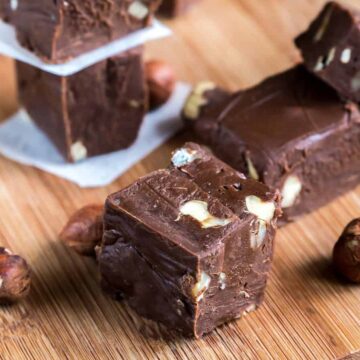 Squares of chocolate fudge with nuts are elegantly arranged on a wooden board, perfect for a cheat day indulgence. Whole hazelnuts are scattered around, adding to the rustic charm. Two fudge squares are stacked neatly with white paper between them for that extra touch of allure.
