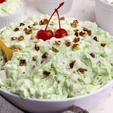 A bowl of green pistachio fluff salad topped with two maraschino cherries and sprinkled with chopped nuts never goes out of style. A wooden spoon is placed in the salad. White bowls containing additional ingredients are in the background. Marshmallows and fabric are in the foreground.