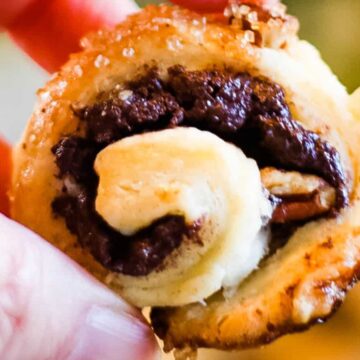 A close-up image shows a hand holding a small, swirled pastry filled with chocolate and pecans. The pastry, which looks like it was crafted in auto draft mode, has a golden-brown, flaky crust with a sprinkle of sugar on top.