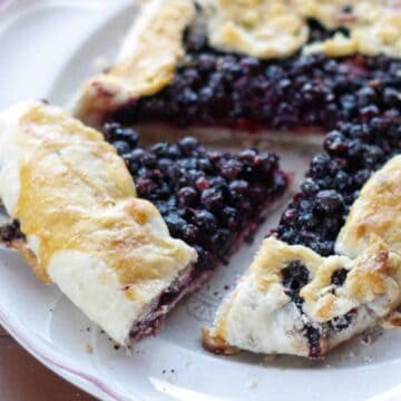 A rustic blueberry galette on a white plate is partially sliced, revealing a flaky, golden-brown crust with a filling of cooked blueberries. Perfect for potluck ideas, this delightful dish sits on a wooden surface.