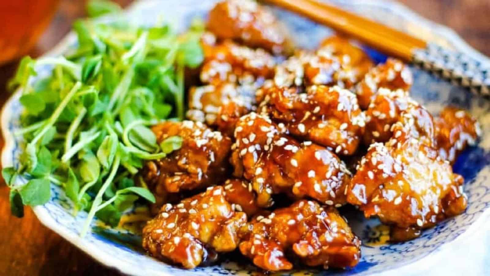 A plate of sesame chicken garnished with sesame seeds sits next to a small portion of fresh green herbs. The dish, swiftly made in an air fryer, appears to have a glossy sauce coating the pieces of chicken, and there are chopsticks resting on the edge of the plate.