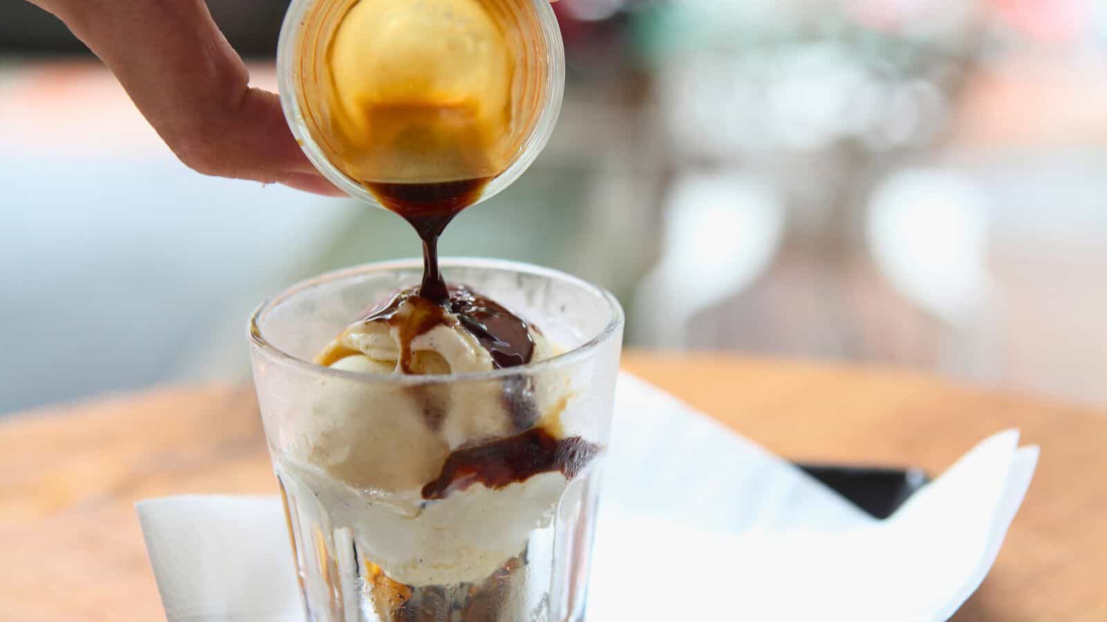 A hand pours a shot of espresso over vanilla ice cream in a glass, creating an affogato. The ice cream is slightly melted, and the espresso is mid-pour, resulting in dark coffee streaks over the pale ice cream. The glass sits on a napkin on a wooden table.