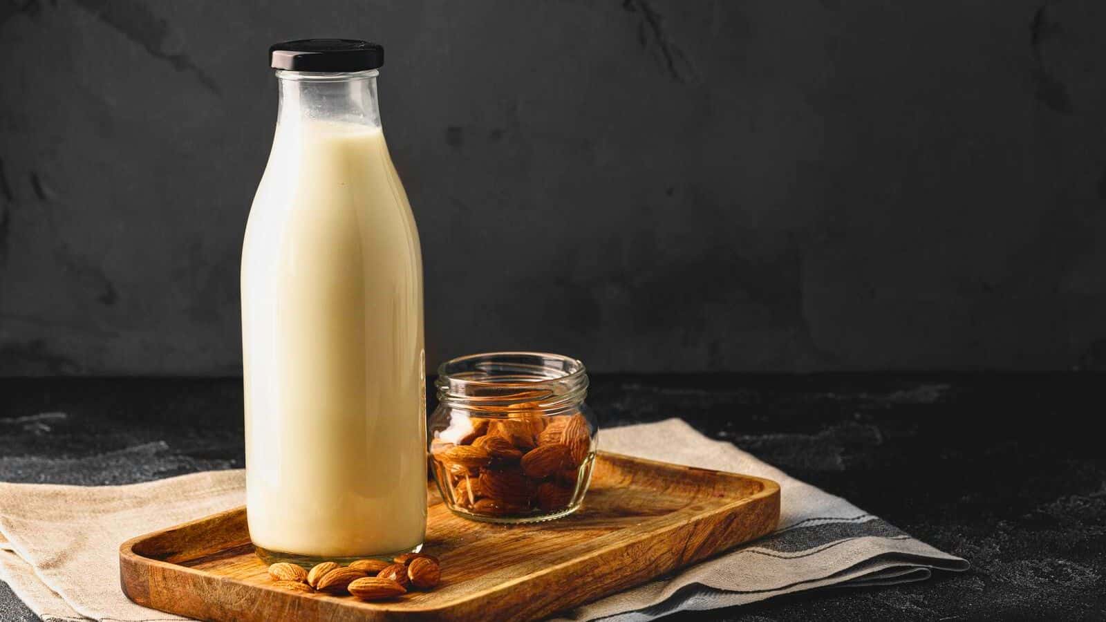 A tall glass bottle of almond milk with a black cap sits on a wooden tray next to a small glass jar filled with almonds. The tray is placed on a folded cloth on a dark, textured surface with a dark background.