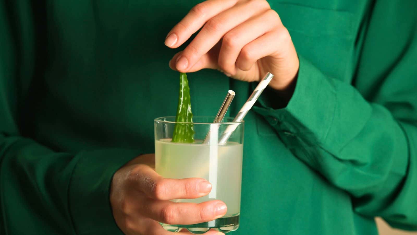 A person in a green shirt is holding a glass of a light green beverage with two striped straws. They are placing or removing a piece of aloe vera into or from the drink. The background is indistinct.