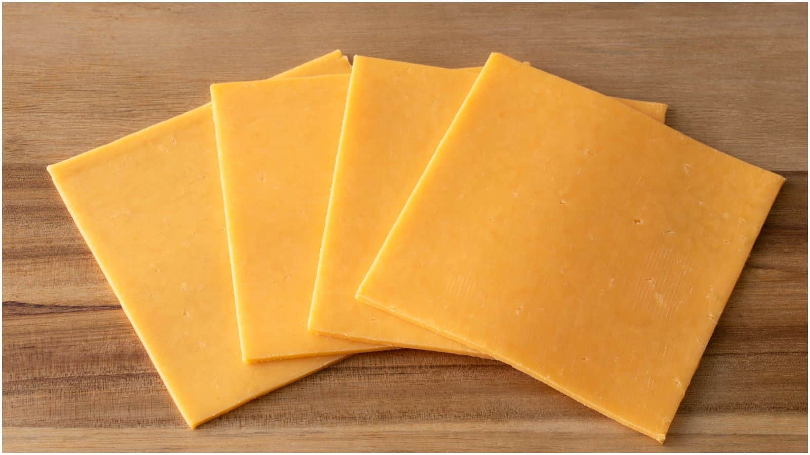 Four square slices of cheddar cheese are arranged in a slightly overlapping pattern on a wooden surface. The cheese has a smooth, uniform texture, and its vibrant orange color contrasts with the brown wood.