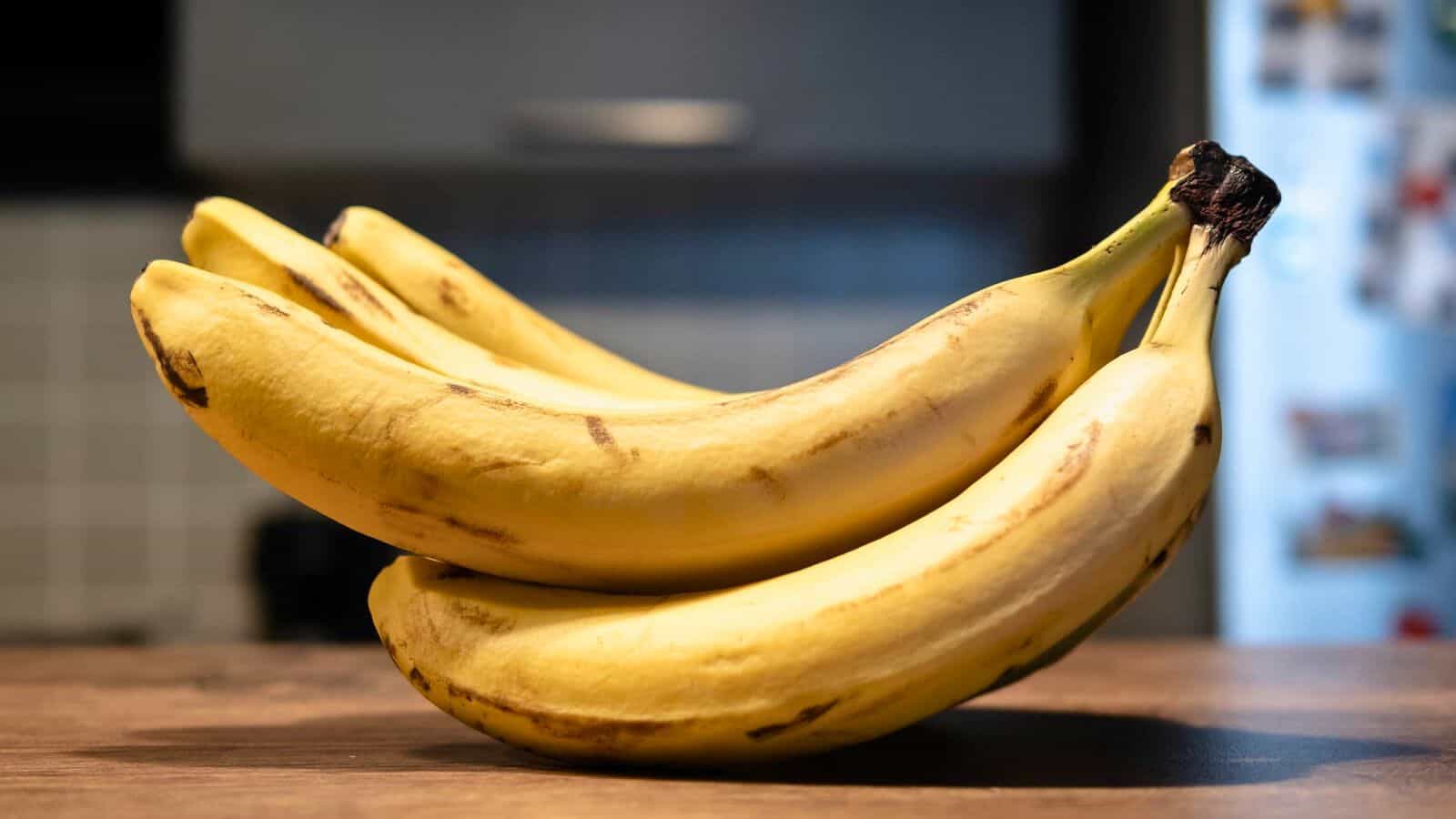 A close-up of a bunch of six ripe bananas with some brown spots, lying on a wooden surface. The background shows a blurred kitchen setting, including a tiled wall and an open refrigerator door.
