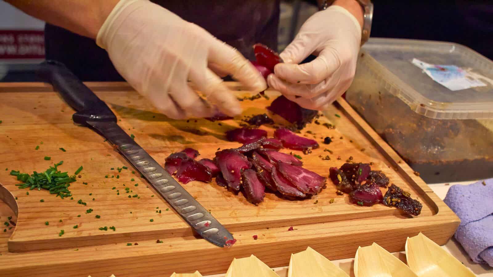 A person wearing gloves slices a piece of cured meat on a wooden cutting board. A knife rests next to the meat, and there are scattered herbs and a container nearby. Disposable paper serving dishes are visible in the foreground.