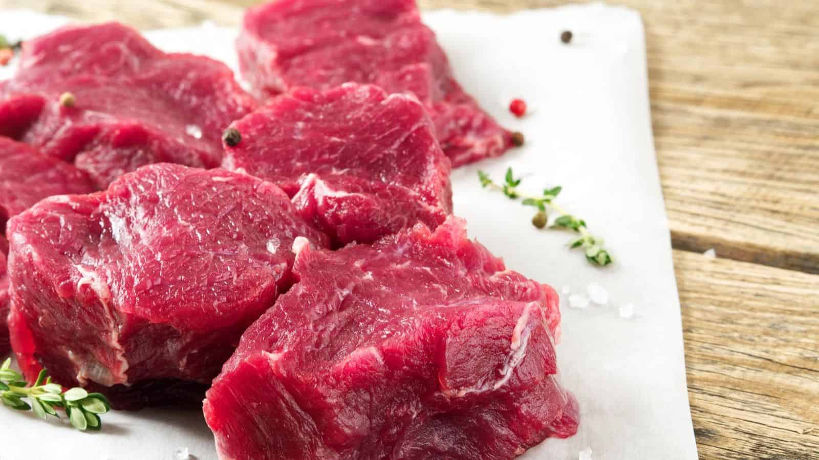 Several raw beef cuts are placed on a white piece of parchment paper, resting on a wooden surface. The meat, representing foods that are becoming too expensive, is garnished with a few sprigs of herbs and scattered peppercorns.