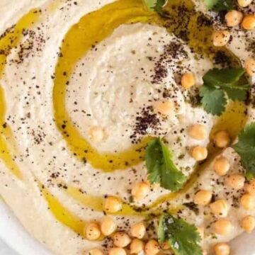 A close-up image of a bowl filled with creamy hummus drizzled with olive oil, garnished with whole chickpeas, fresh cilantro leaves, and a sprinkle of ground spices. This delicious dip, ideal for Rosh Hashanah recipes, is placed on a white marble surface.
