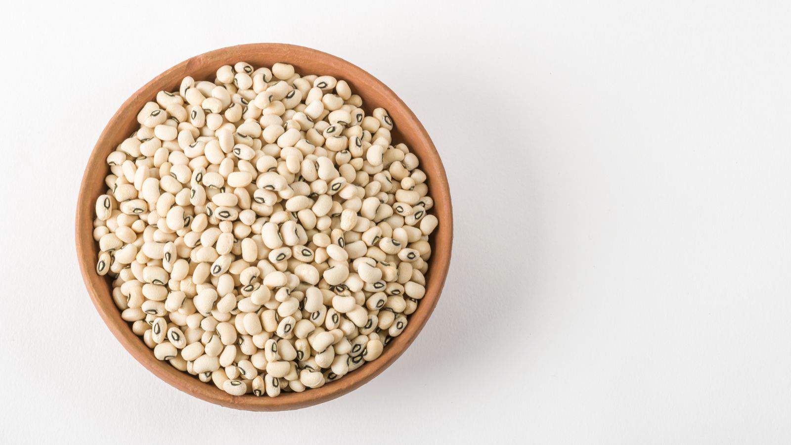 A wooden bowl filled with uncooked black-eyed peas is placed on a plain white surface. The peas are light-colored with characteristic small black spots.